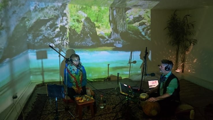SON tocando en vivo: Viky cantando y Agustín en el udu, con un río proyectado sobre la pared